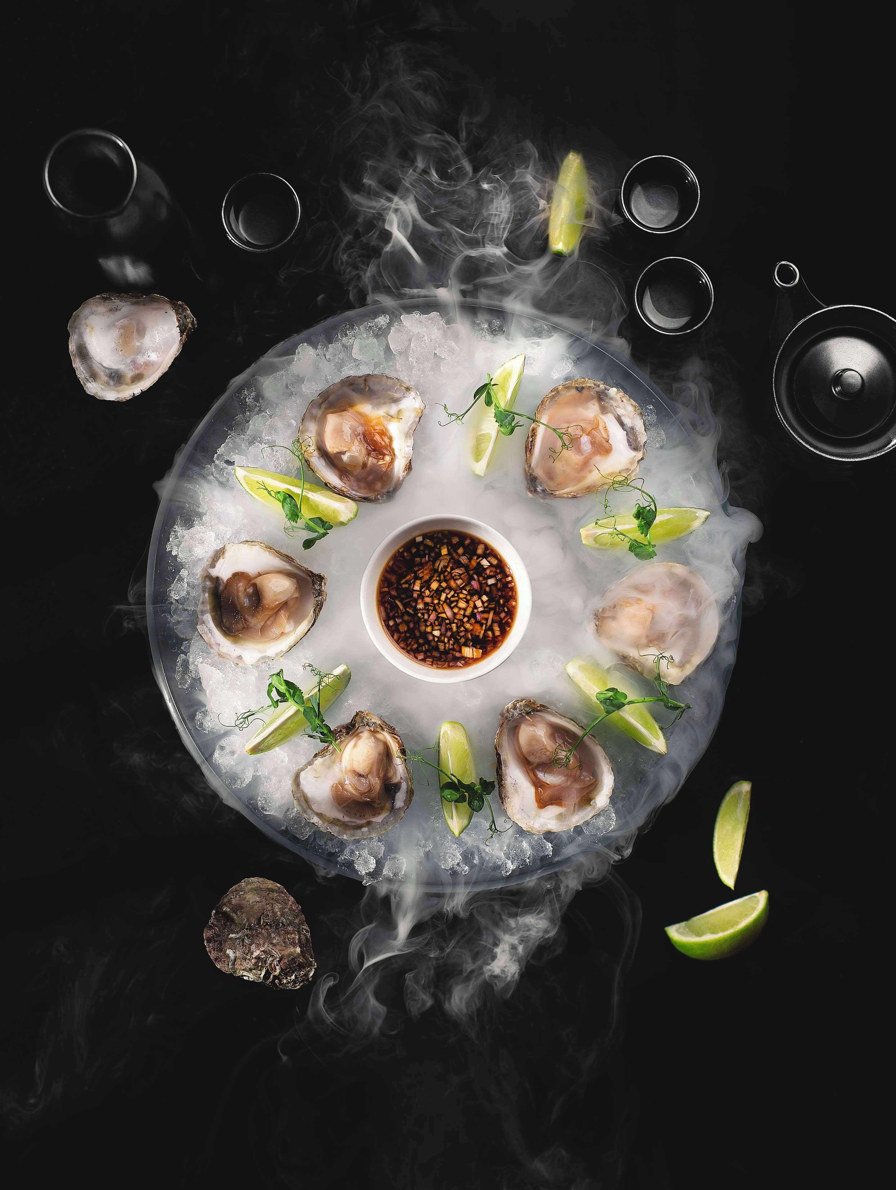 A platter of oysters on ice with a dipping sauce, garnished with herbs and lime slices.