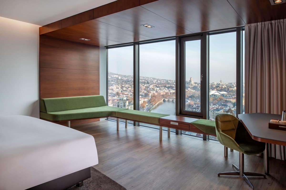 A modern hotel room with a large window offering a city view, a green sofa, a desk, and a bed.