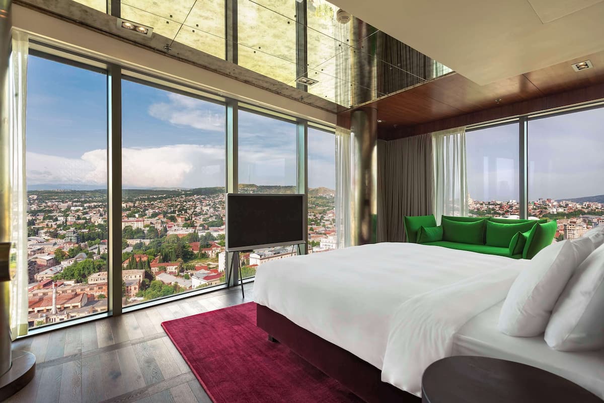A modern hotel room with a large bed, a TV, and a view of a cityscape through floor-to-ceiling windows.