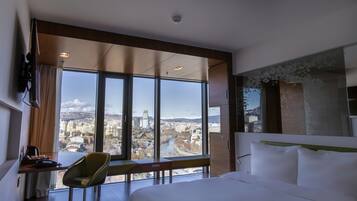 Una habitació d'hotel amb un llit gran, un escriptori amb una cadira i vista a un paisatge urbà a través de grans finestres.
