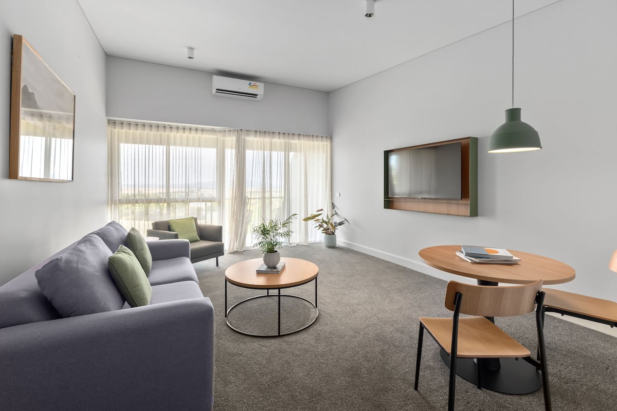 A modern living room with a grey sofa, a round coffee table, and a dining table with chairs. There is a potted plant on the coffee table and a large mirror on the wall.