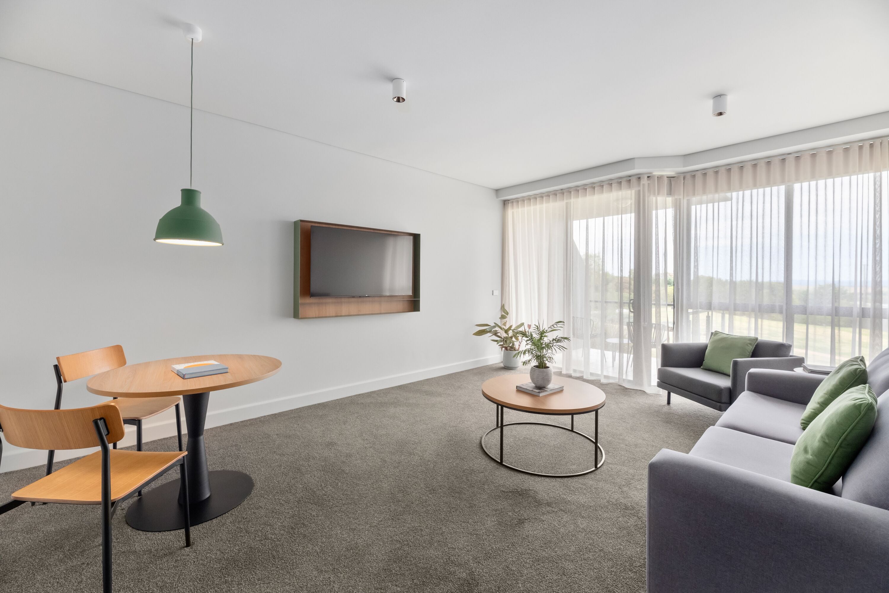 Un salón moderno con una mesa redonda de madera, dos sillas, un sofá gris con cojines verdes, una lámpara colgante verde, un televisor de pantalla plana montado en la pared y una planta en maceta sobre una mesita redonda.