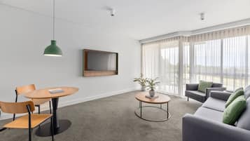 Un salón moderno con una mesa redonda de madera, dos sillas, un sofá gris con cojines verdes, una lámpara colgante verde, un televisor de pantalla plana montado en la pared y una planta en maceta sobre una mesita redonda.