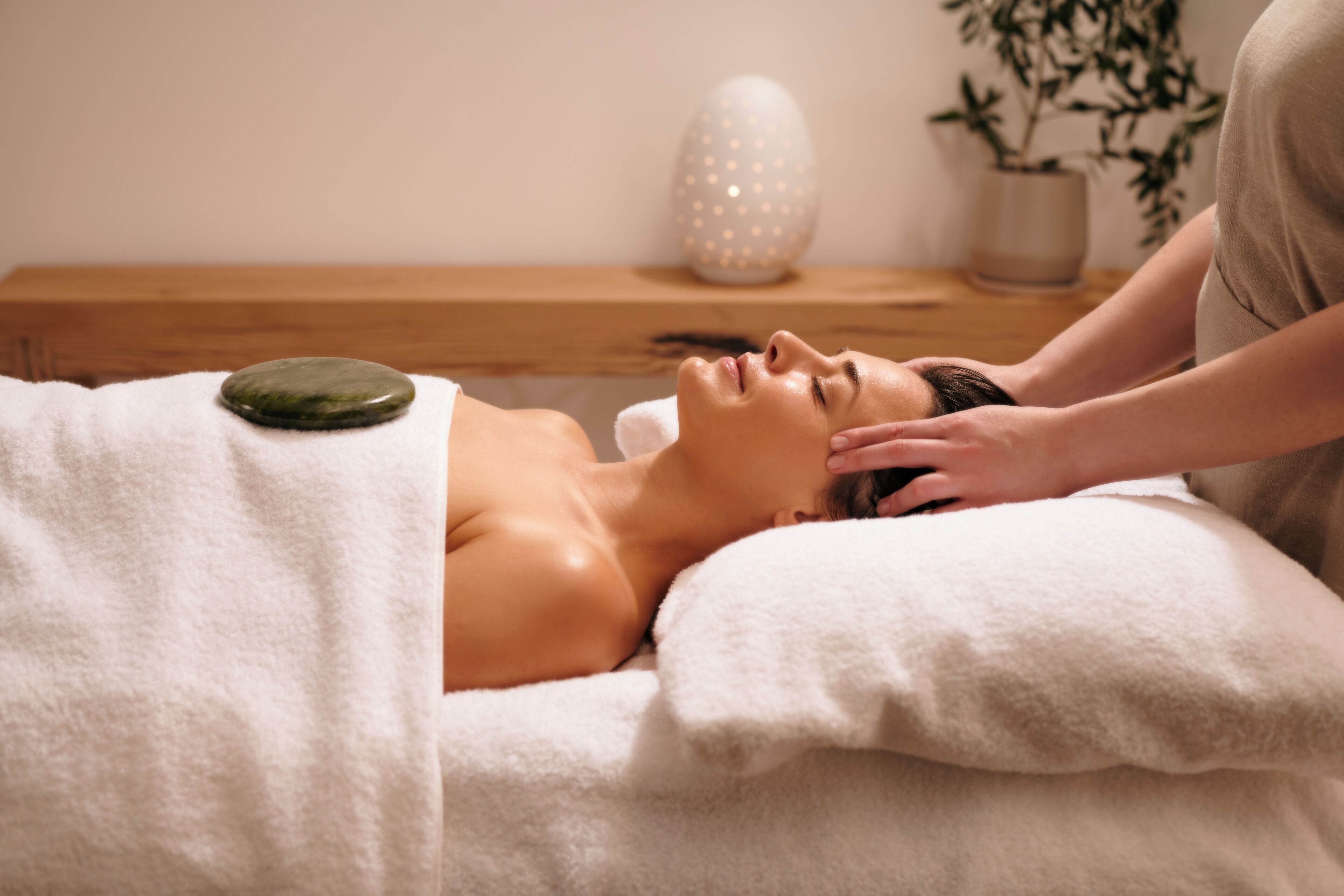 A person receiving a massage with a stone on their back, a white towel, and a plant in the background.