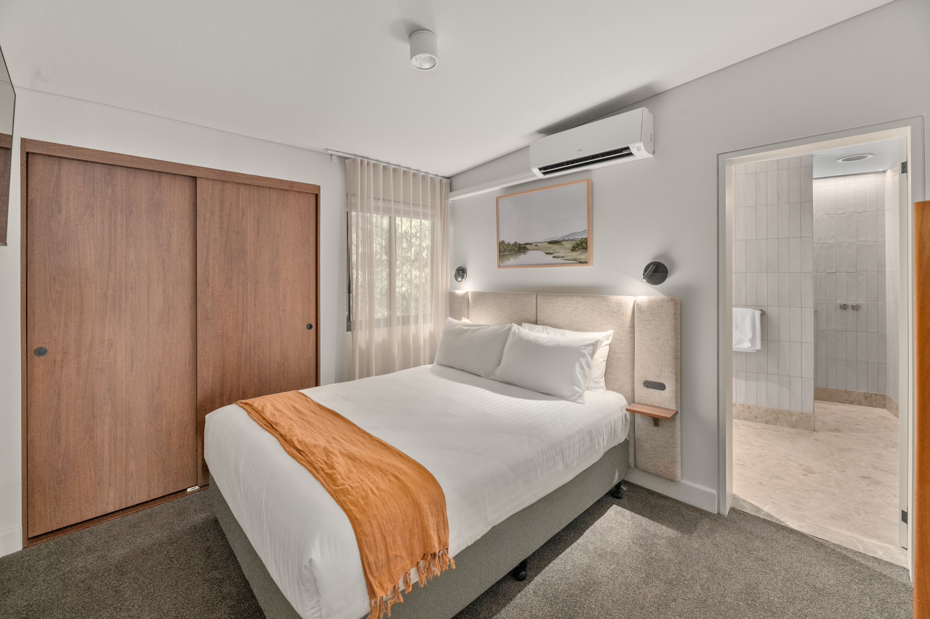 A modern bedroom with a large bed, a wooden wardrobe, an air conditioning unit, a framed picture on the wall, and a shower area.