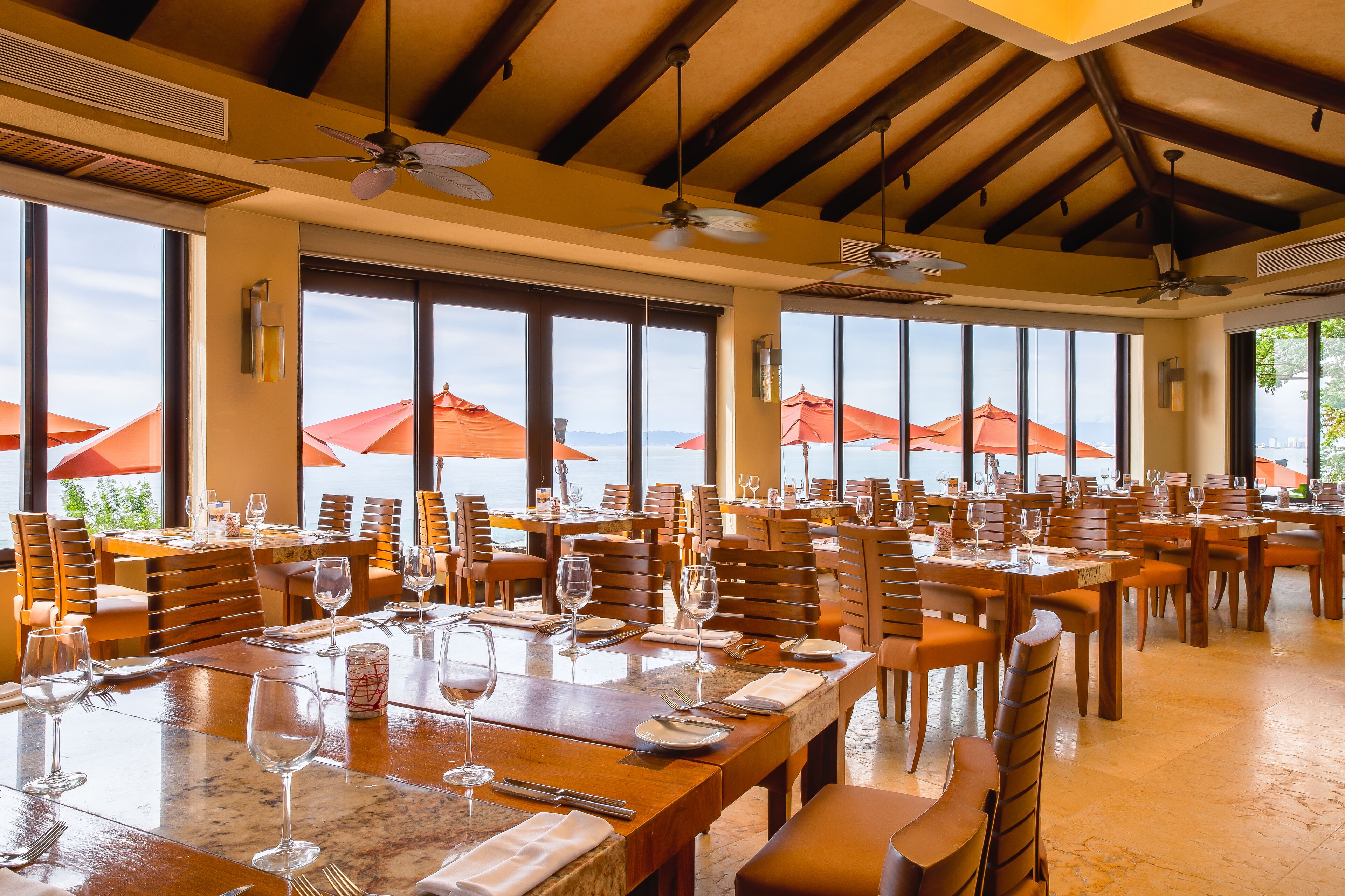 A spacious dining area with multiple tables and chairs, large windows with red umbrellas, and ceiling fans.