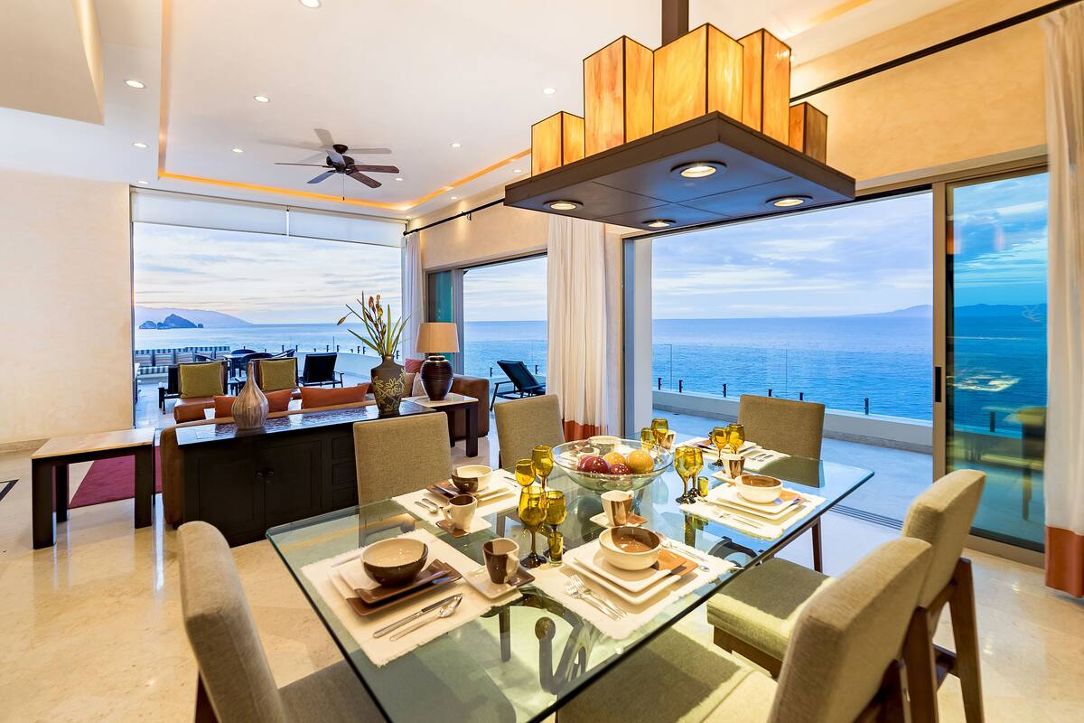 A modern dining area with a glass table, chairs, and a view of the ocean.