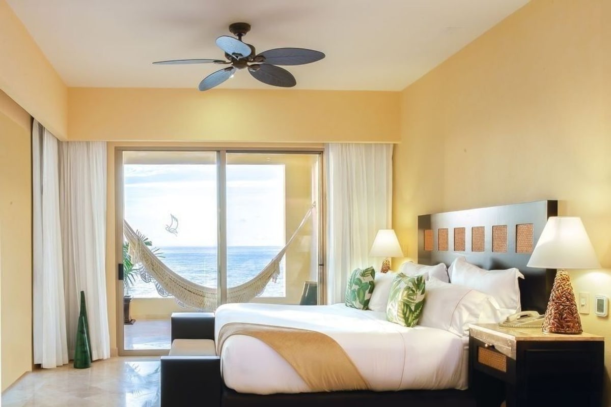 Una habitación de hotel con cama, mesita de noche, lámpara, ventilador de techo y vistas al océano, además de una hamaca.