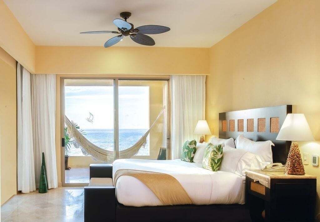 Una habitación de hotel con cama, mesita de noche, lámpara, ventilador de techo y vistas al océano, además de una hamaca.