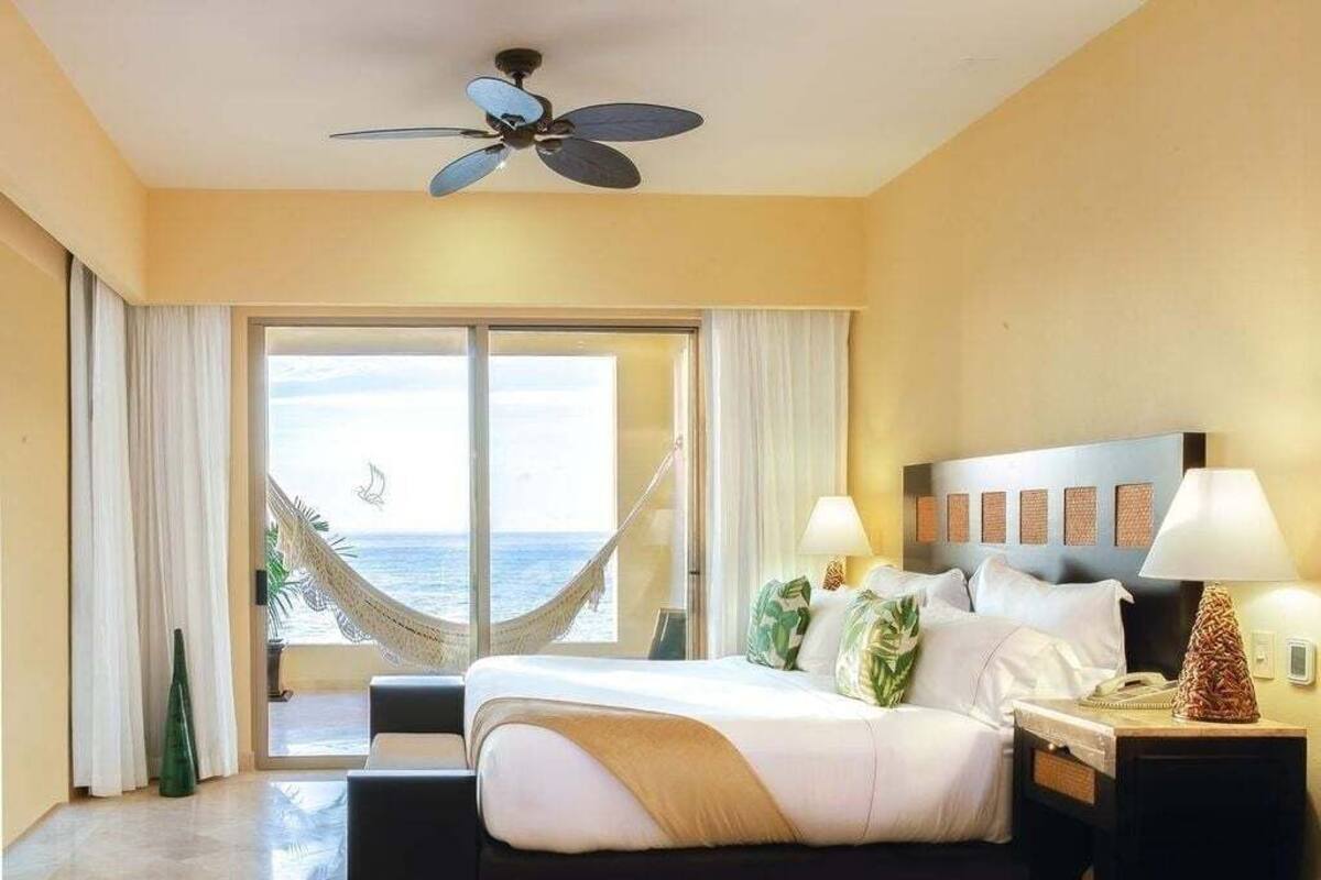 A hotel room with a bed, a nightstand, a lamp, a ceiling fan, a view of the ocean, and a hammock.