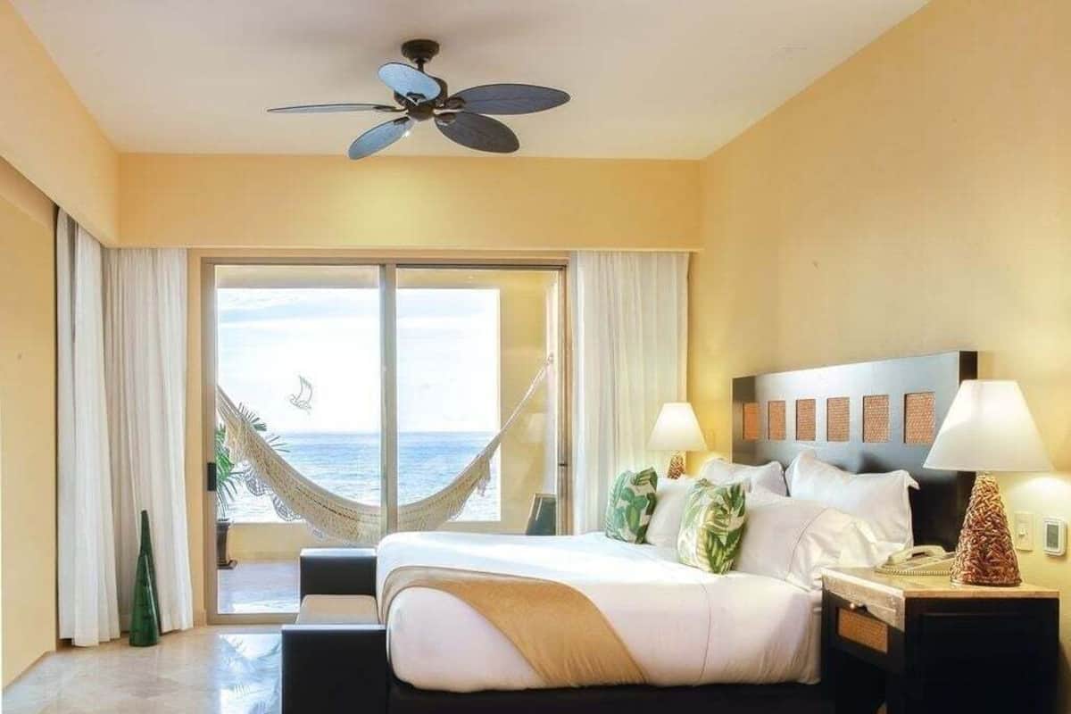A hotel room with a bed, a nightstand, a lamp, a ceiling fan, a view of the ocean, and a hammock.
