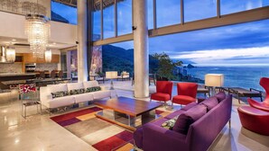 A spacious lobby with a large seating area, a kitchen in the background, and a view of the sea and mountains through large windows.