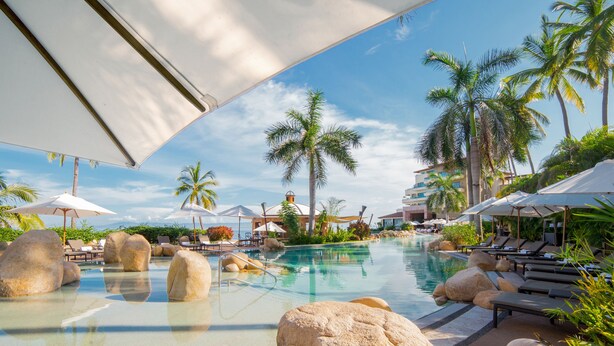 A resort pool area with lounge chairs, umbrellas, and palm trees.
