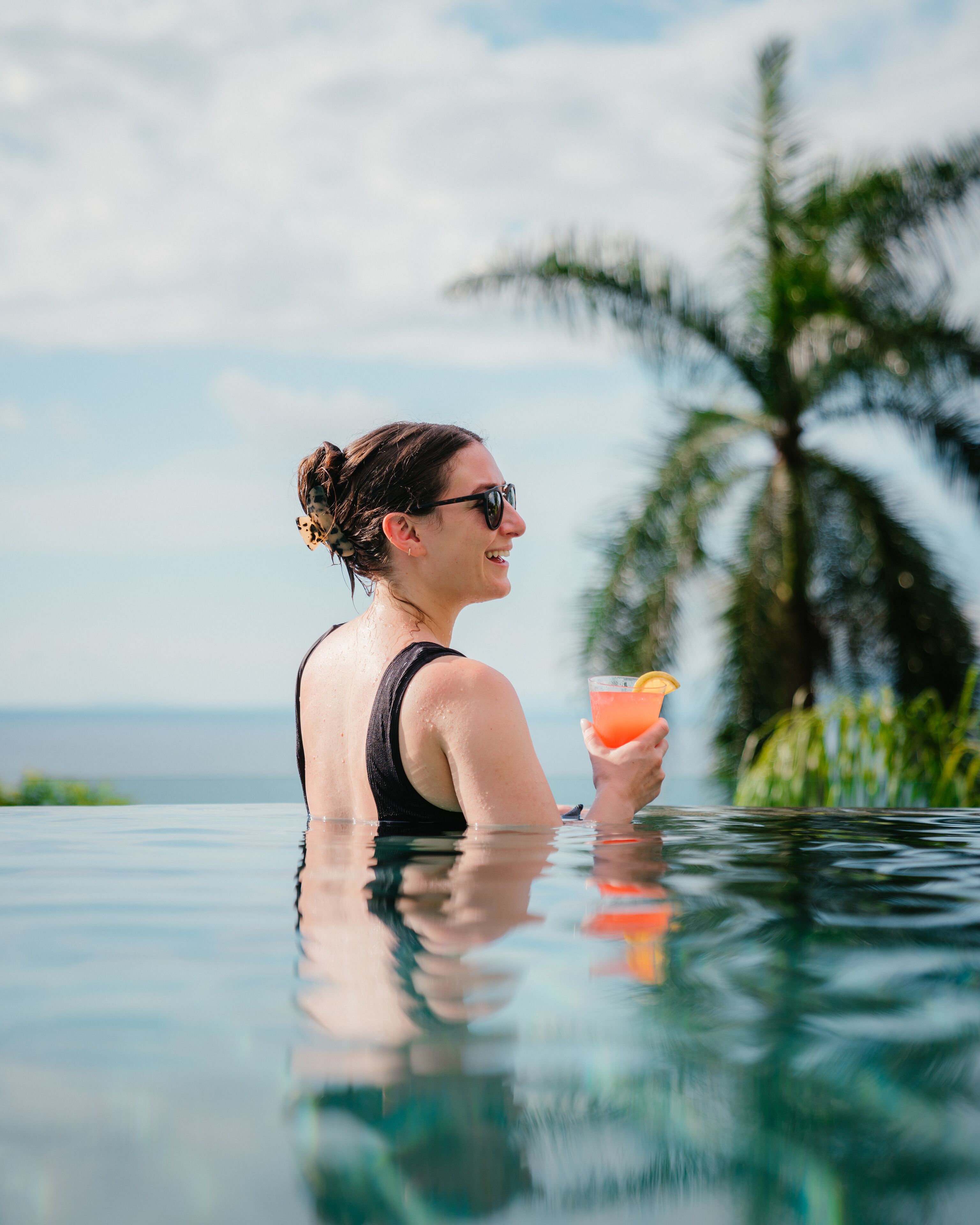 Une personne dans une piscine, tenant un verre à la main, avec une vue dégagée sur le ciel et des palmiers en arrière-plan.