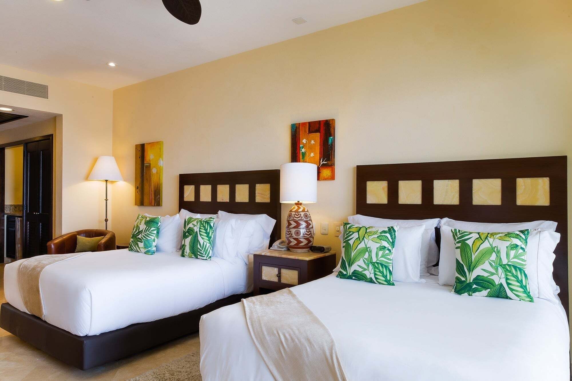 Habitación de hotel con dos camas, una mesita de noche con lámpara, un cuadro en la pared y una puerta.