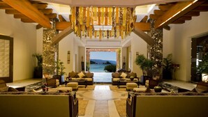 A spacious lobby with a view of the sea, featuring a central seating area with sofas and armchairs, a stone wall, and a wooden ceiling structure.