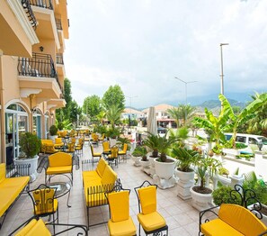 An outdoor seating area with yellow chairs and tables, surrounded by potted plants and a view of the sea.