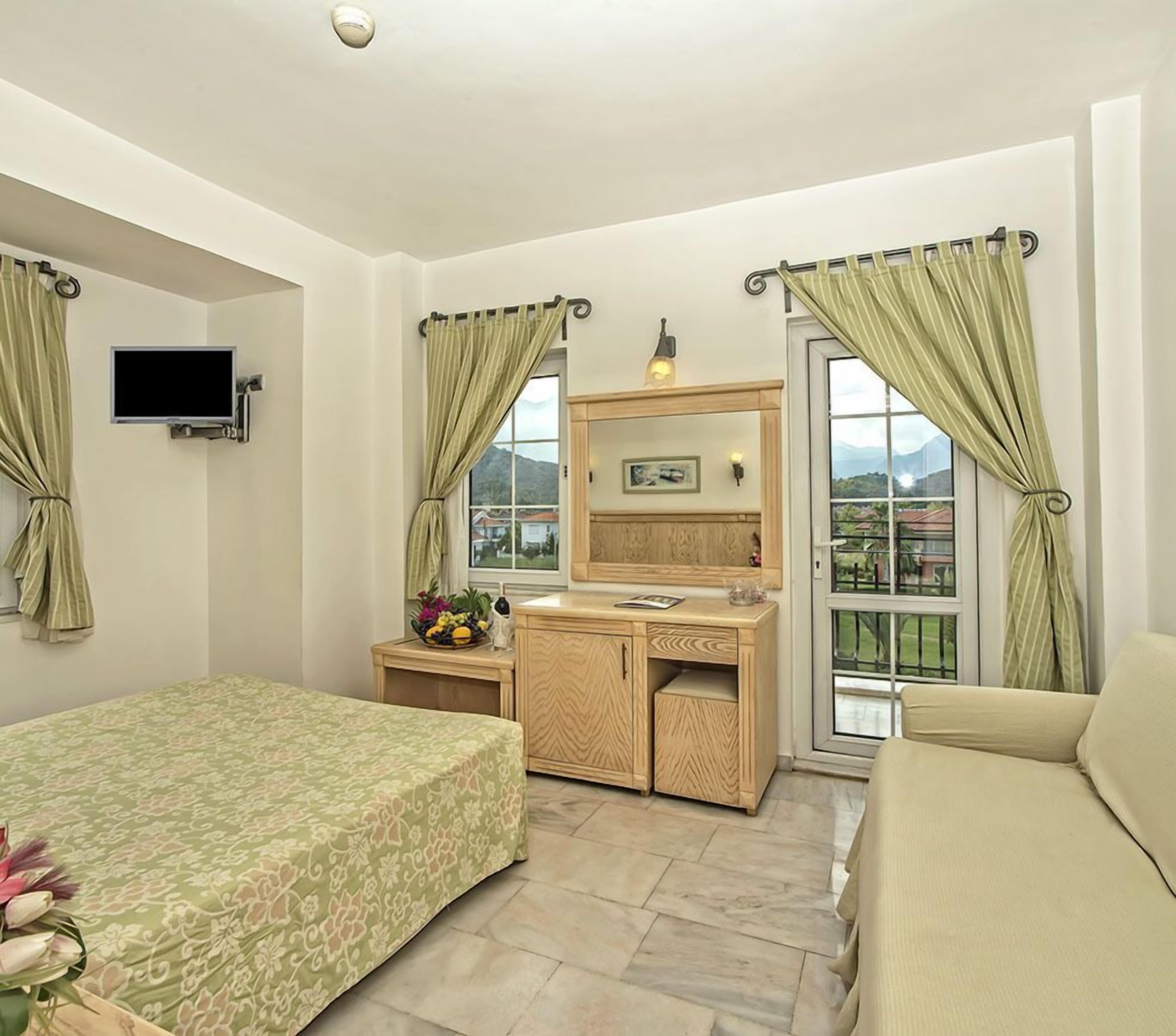 Ein Schlafzimmer mit einem Bett, einem Schreibtisch, einem Sessel, einem Fernseher und einem Fenster mit Vorhängen.