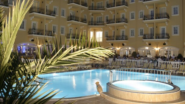 Ein großer, überdachter Swimmingpool, umgeben von Balkonen mit Geländern, Palmen und einer Sitzgelegenheit mit Stühlen.