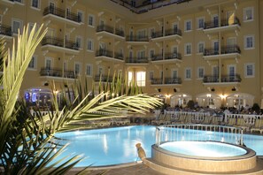 Una amplia piscina cubierta rodeada de balcones con barandillas, palmeras y una zona de estar con sillas.