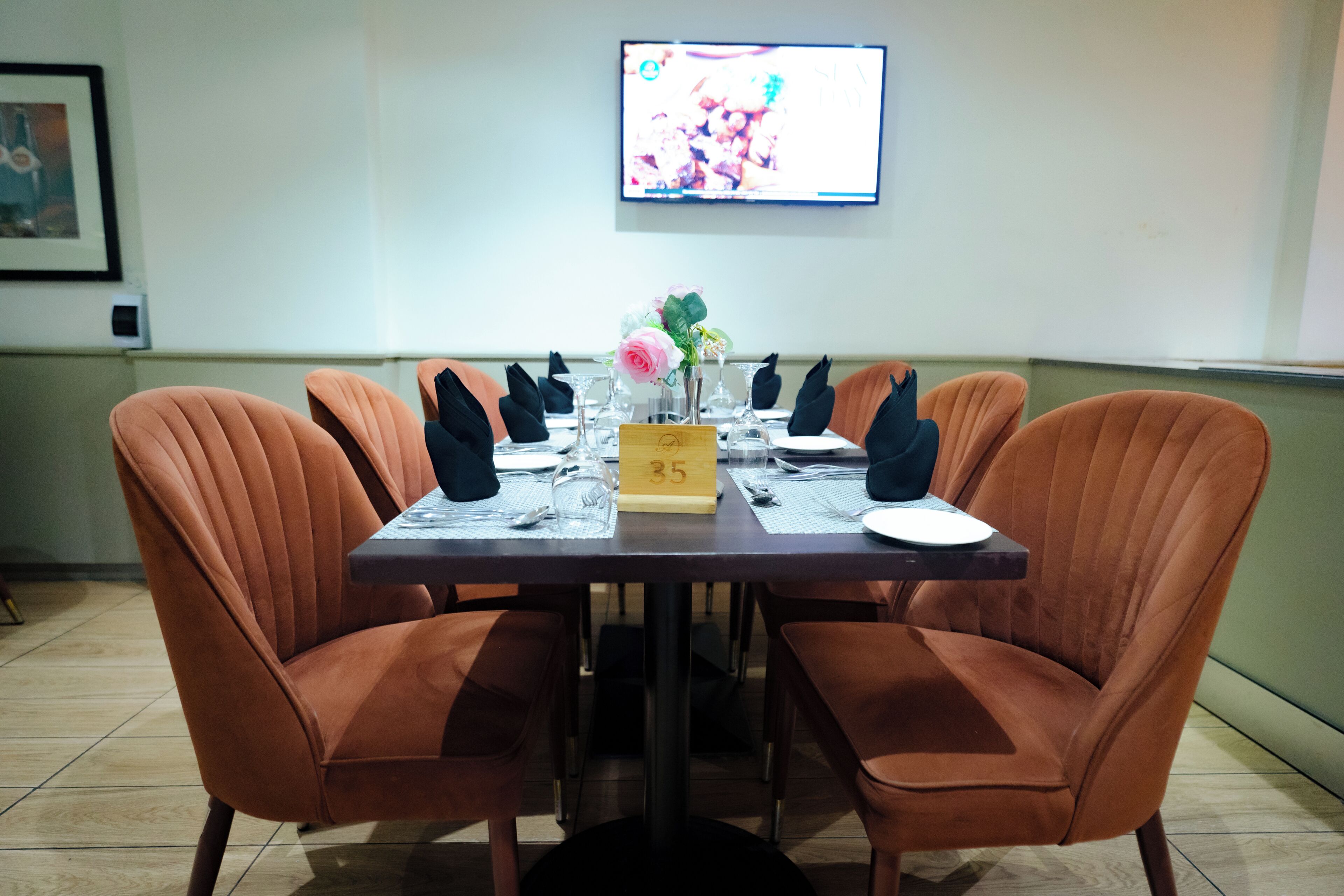A dining table with numbered place settings, orange upholstered chairs, and a television mounted on the wall.