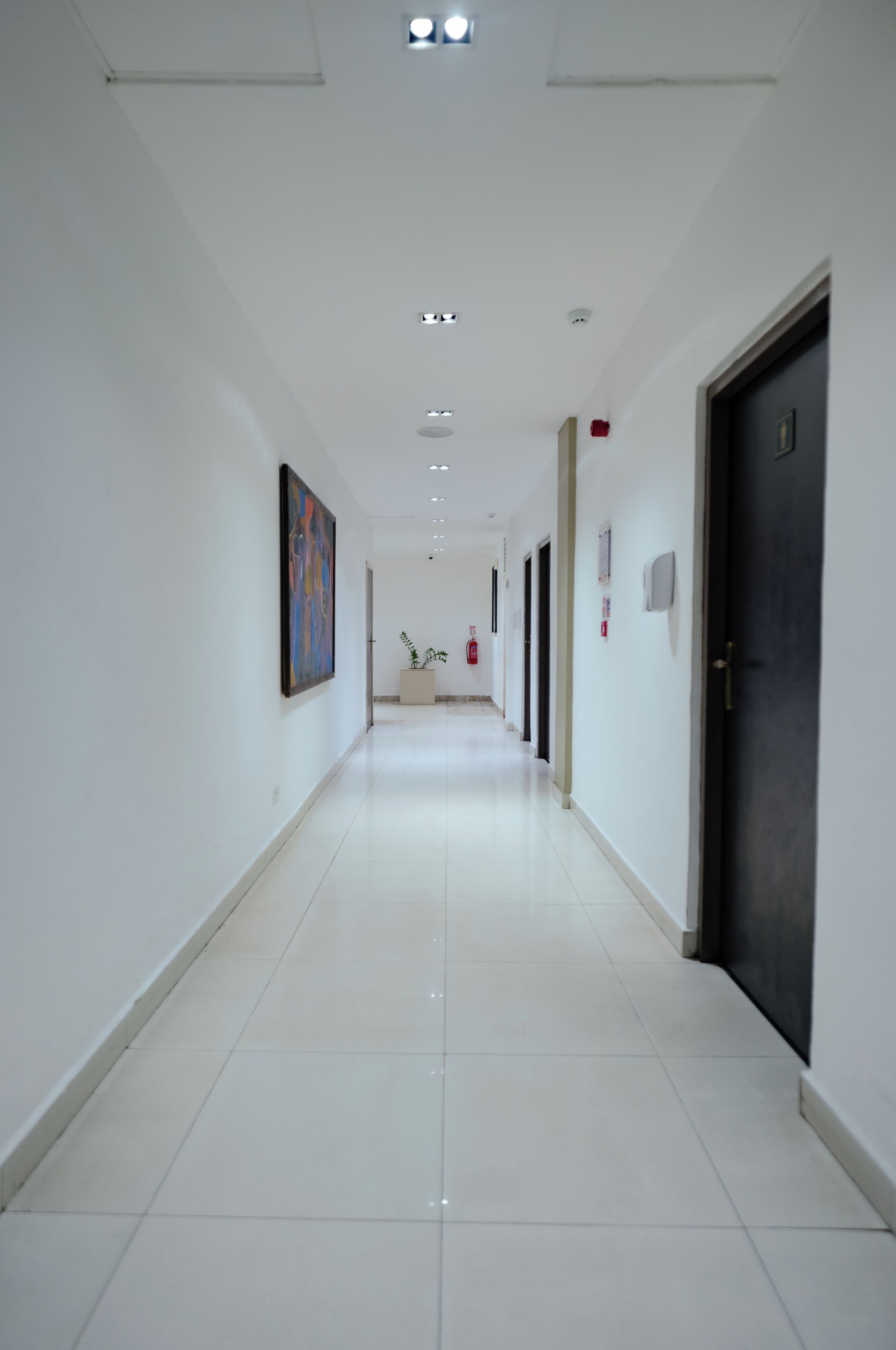 A long, narrow hallway with white walls, a tiled floor, and doors on one side.