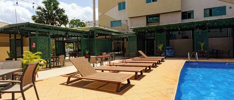 Zona de piscina de hotel con tumbonas, piscina y mobiliario exterior.