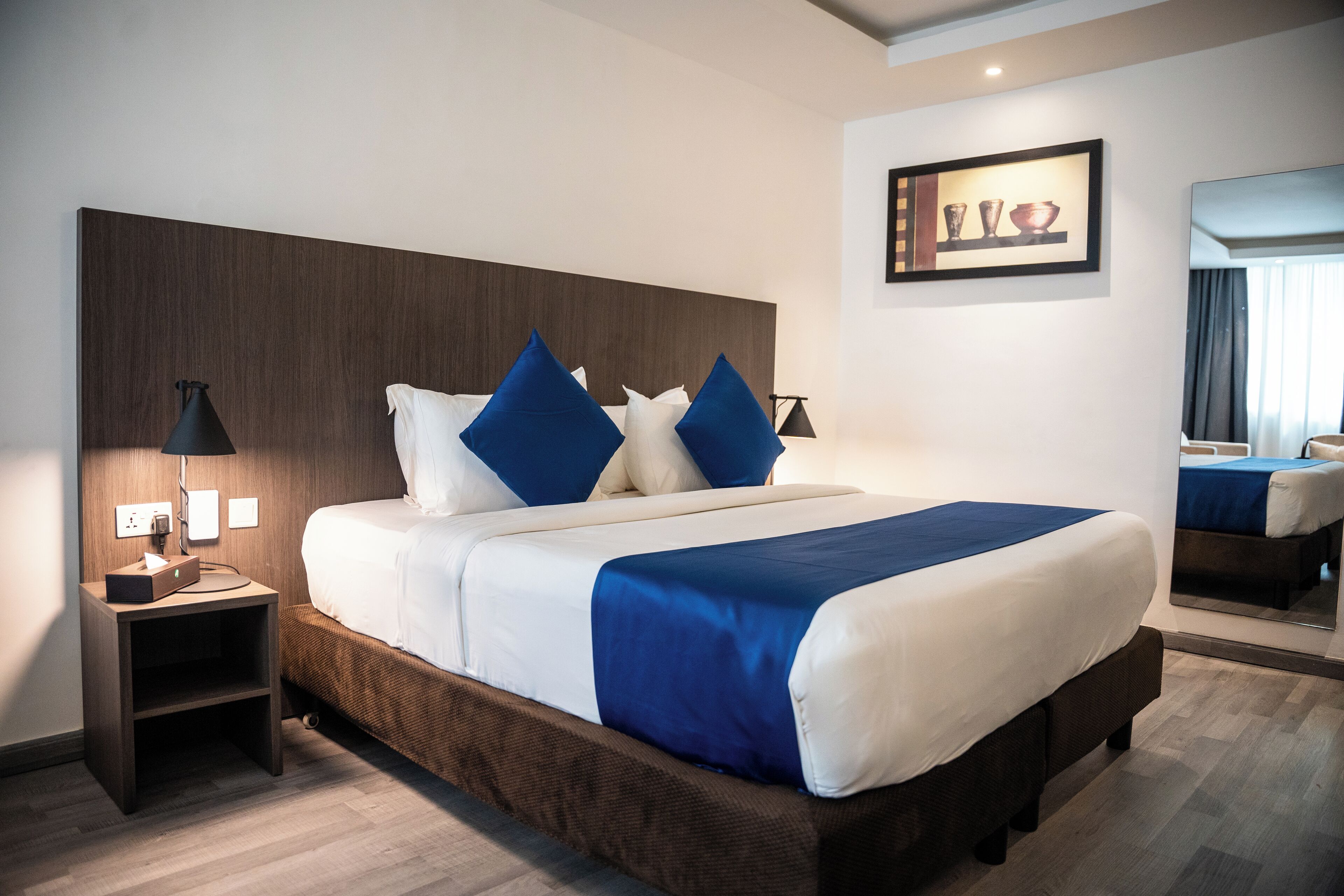 A modern hotel room with a large bed, a nightstand with a lamp, a framed picture on the wall, and a mirror reflecting the room.
