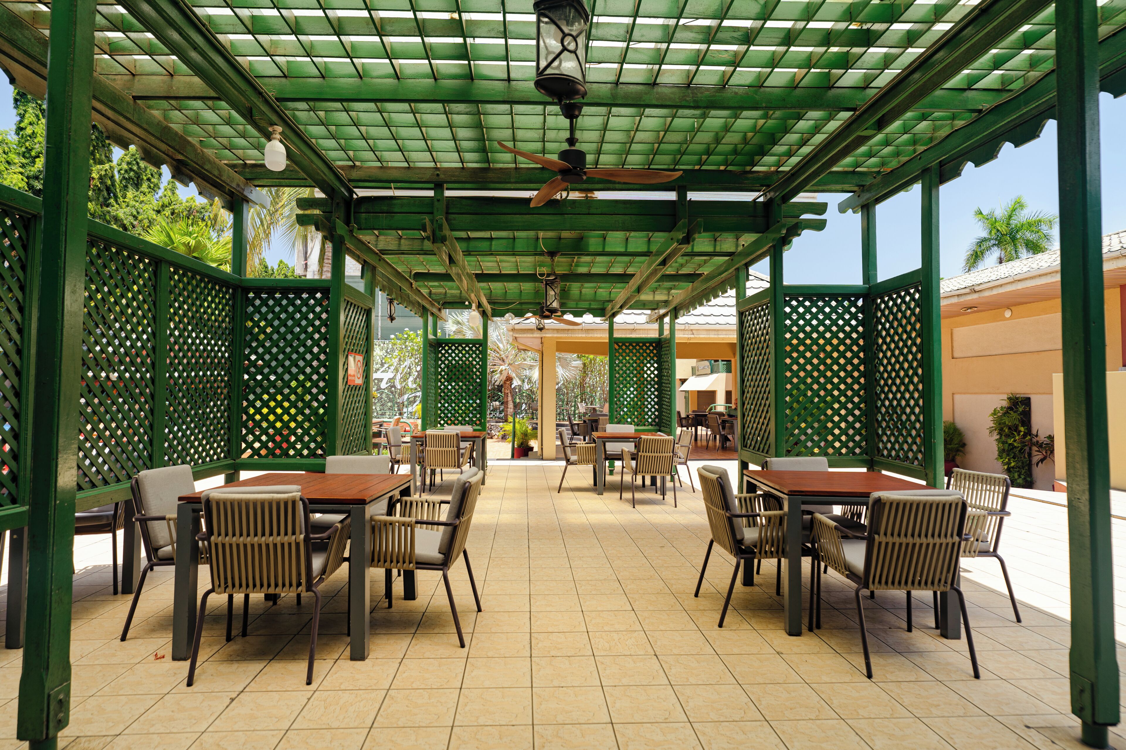 Un espacio exterior techado para comer con mesas y sillas, un ventilador de techo y vistas del entorno.