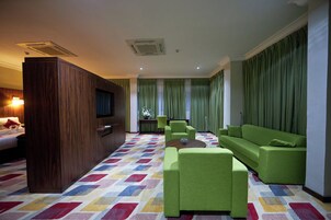 A hotel room with a large bed, a TV, a green sofa, a green armchair, a small table, and a colorful carpet.