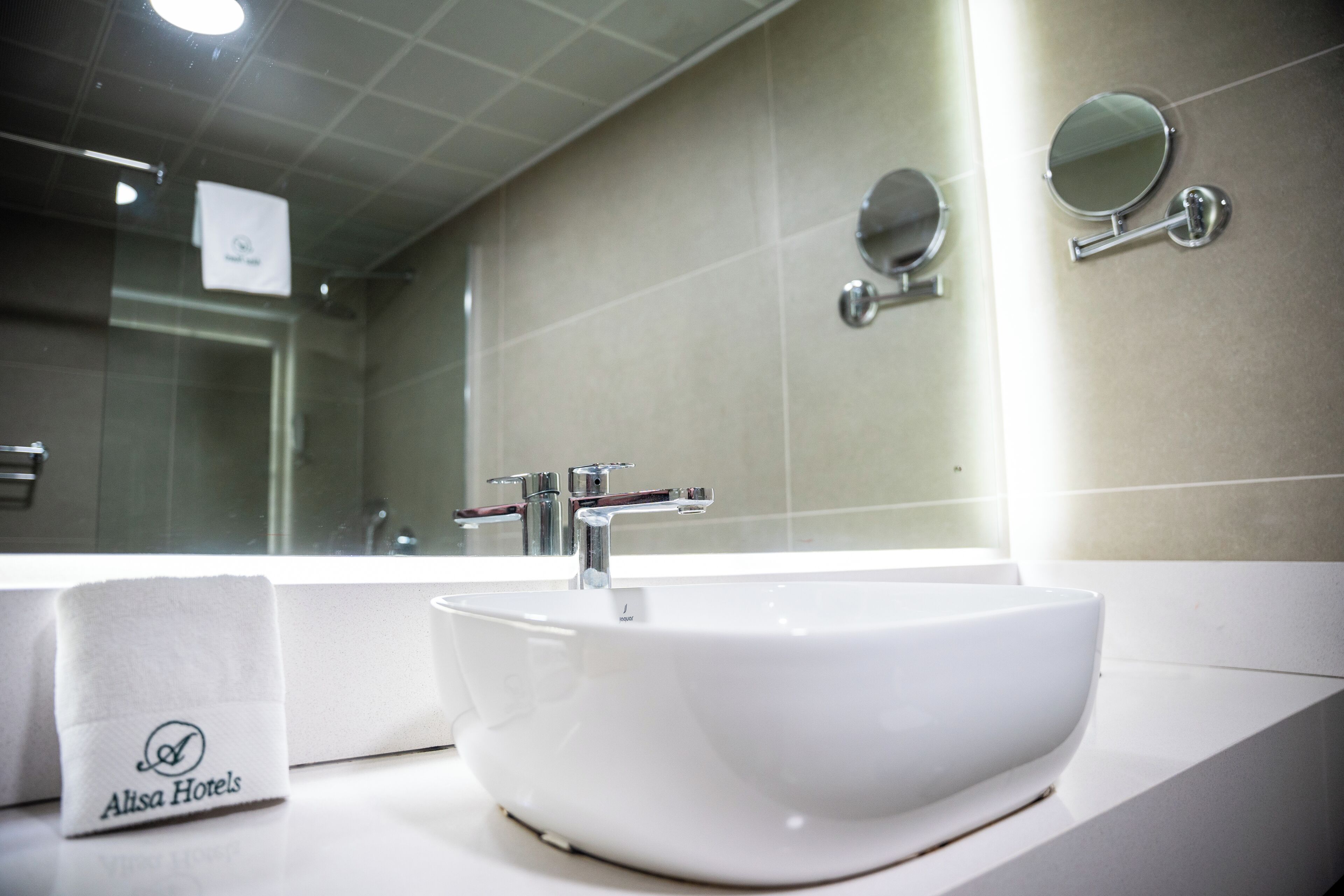 Un baño moderno con un lavamanos blanco, dos espejos y un perchero con la etiqueta 'Alisa Hotels'.