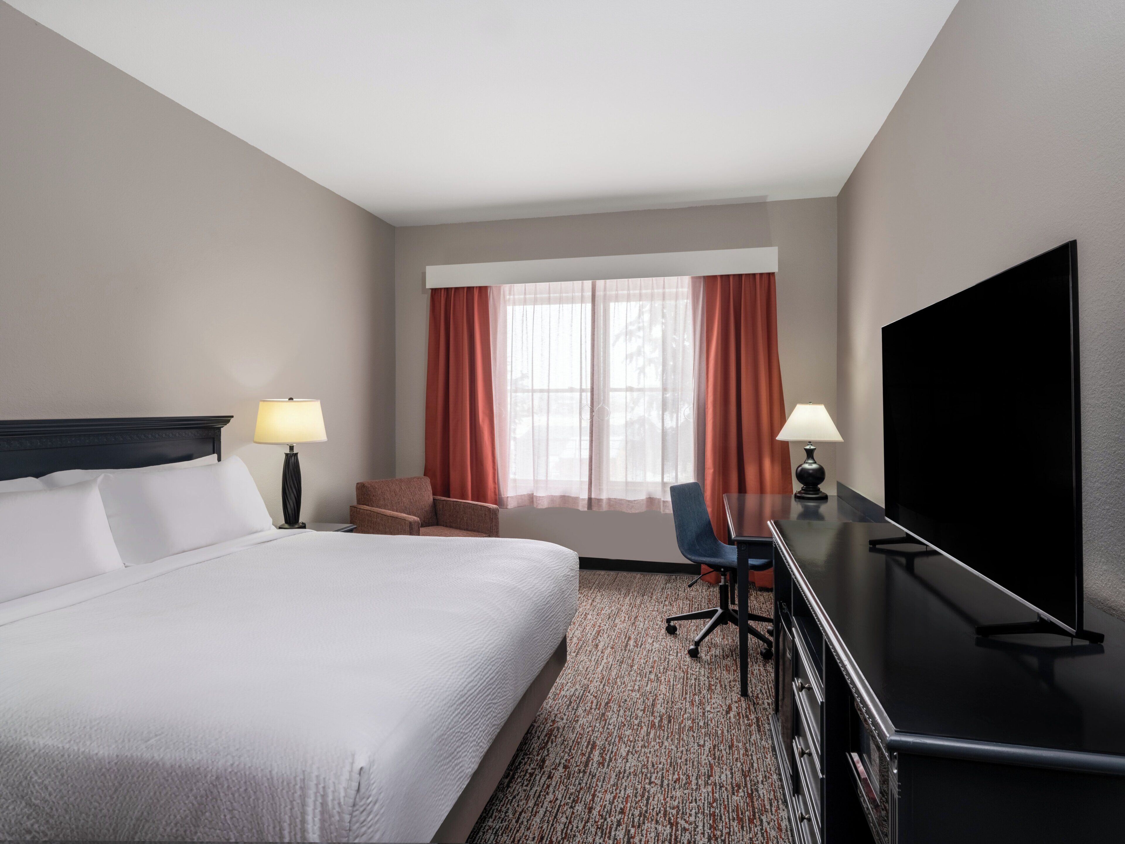 A hotel room with a large bed, a desk with a flat-screen TV, two chairs, a lamp, and a window with curtains.