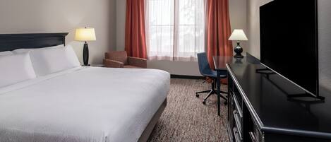 A hotel room with a large bed, a desk with a flat-screen TV, two chairs, a lamp, and a window with curtains.