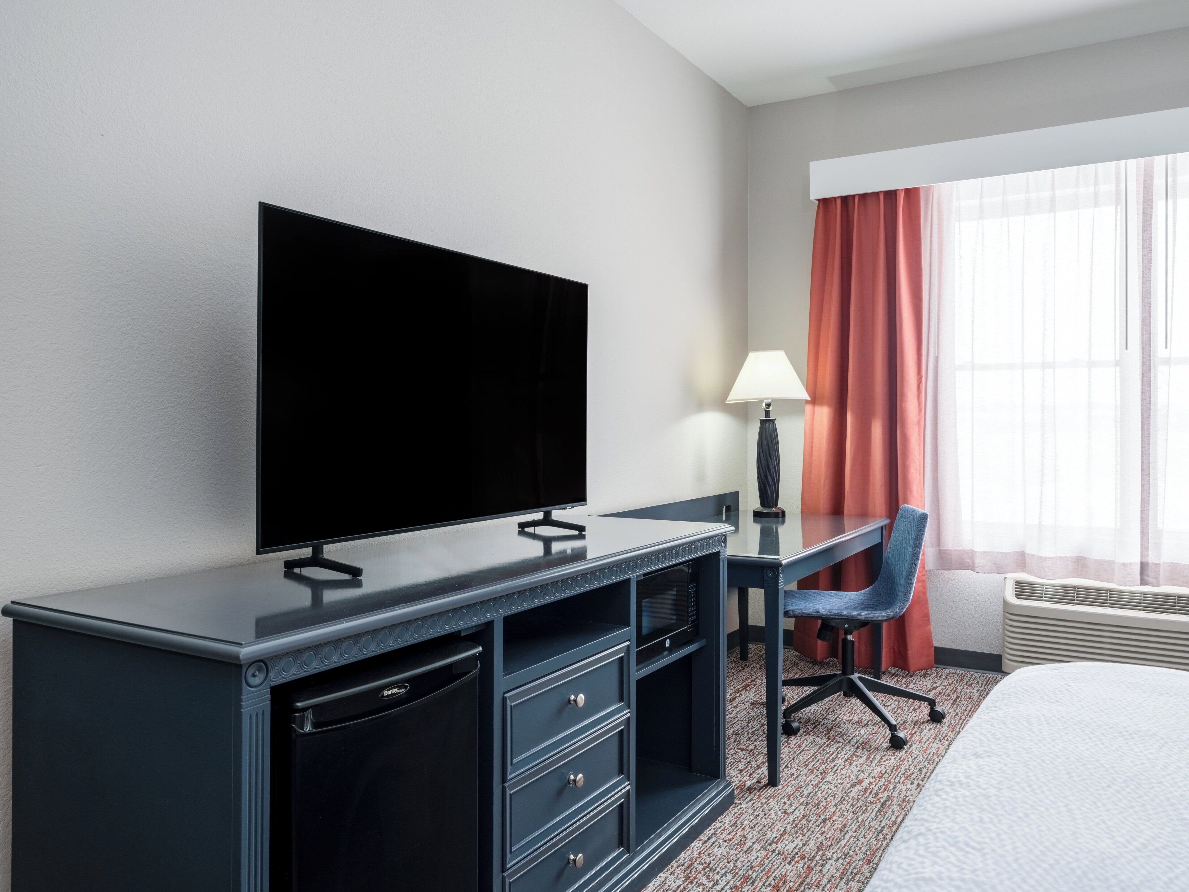 Habitación de hotel con televisión de pantalla plana, escritorio con lámpara, una silla, una cama y ventana con cortinas.