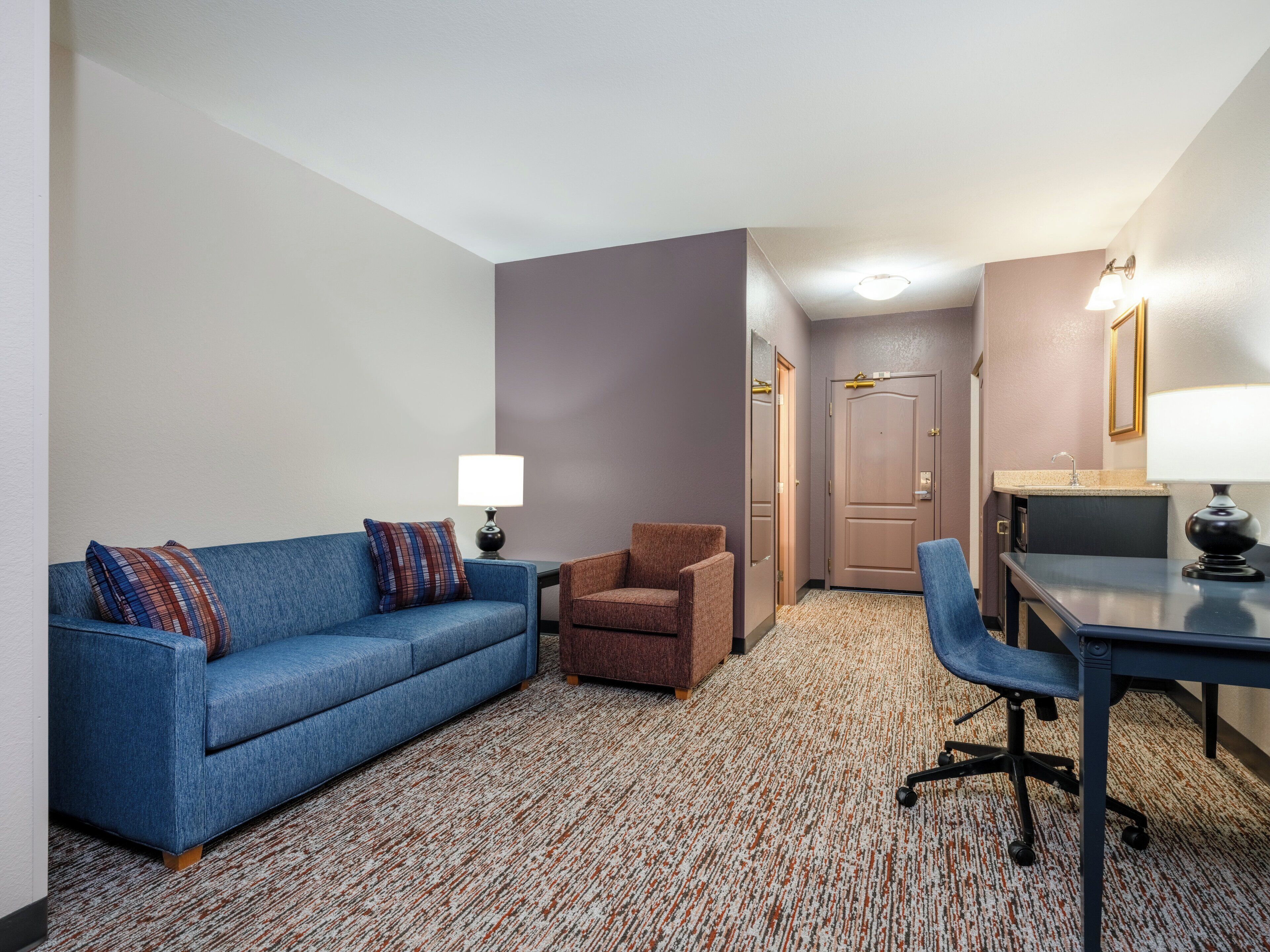 Habitación de hotel con sofá azul, sillón marrón, escritorio con lámpara y suelo alfombrado.