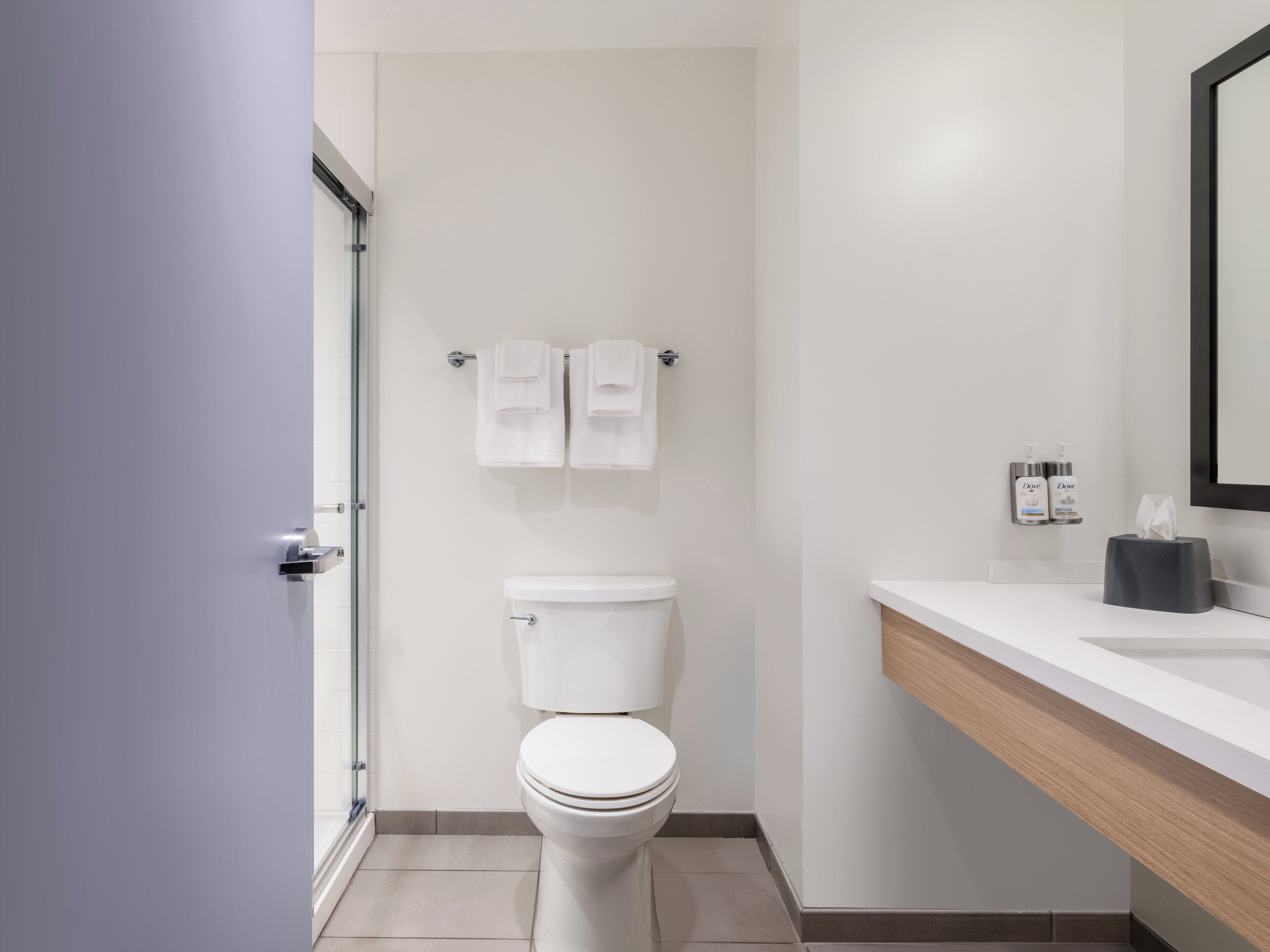 Un baño moderno con inodoro, toallero con dos toallas, lavamanos con dispensador de jabón y espejo.