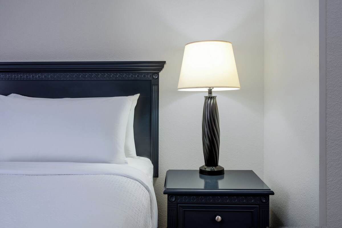 Una cama bien hecha con sábanas y almohadones blancos, una mesita de noche de madera oscura con una lámpara, y una pared con textura.