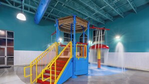 Un espace de jeux aquatiques intérieur avec une structure de jeu bleue et jaune, une fontaine et un sol carrelé.