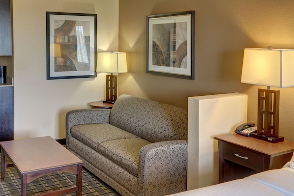 Habitación de hotel con sofá, una mesita, una mesita de noche con lámpara, teléfono y un cuadro en la pared.