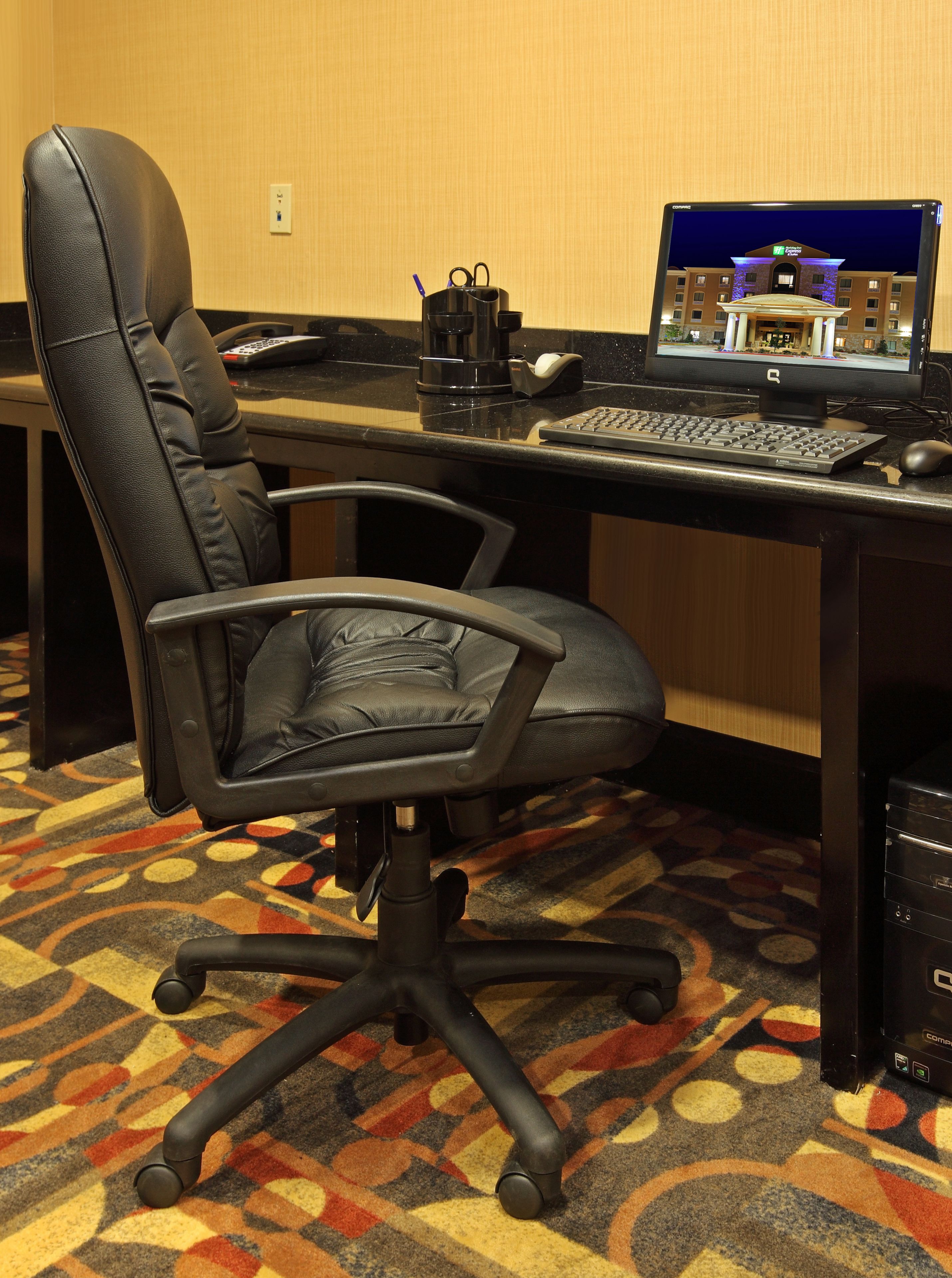Habitación de hotel con una silla de oficina de cuero negro, un escritorio con computadora, teclado, ratón y una cafetera.