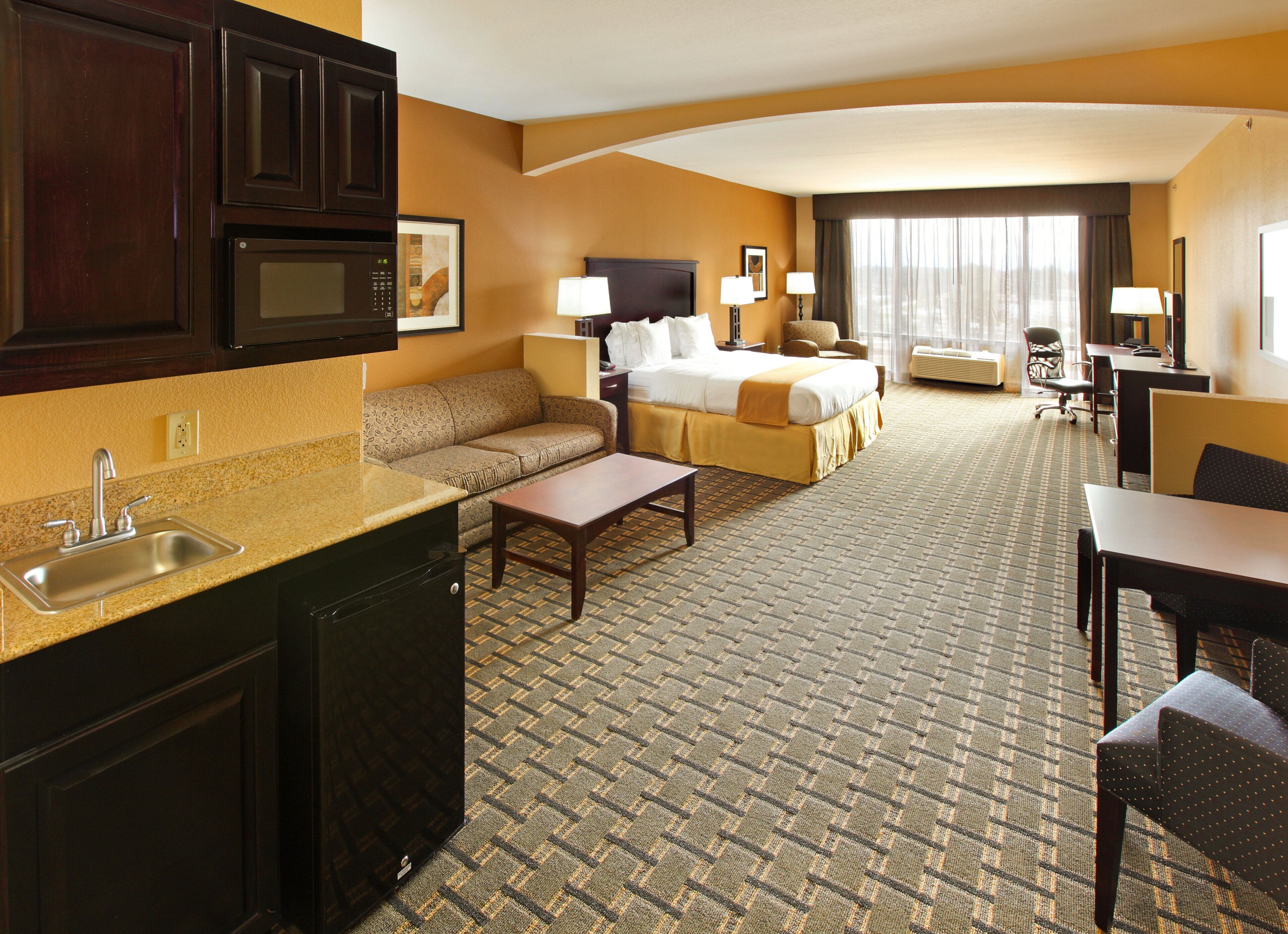 A hotel room with a kitchenette, a sofa, a bed, a desk, and a chair.