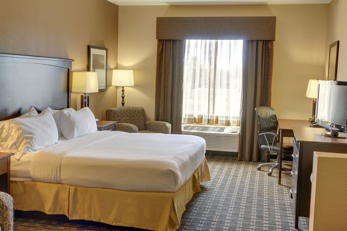 Een hotelkamer met een groot bed, een bureau met een computer, een stoel, een klein tafeltje en een raam met gordijnen.