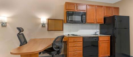 A compact kitchen with wooden cabinets, a black microwave, a black refrigerator, and a dining area with a wooden table and black office chair.