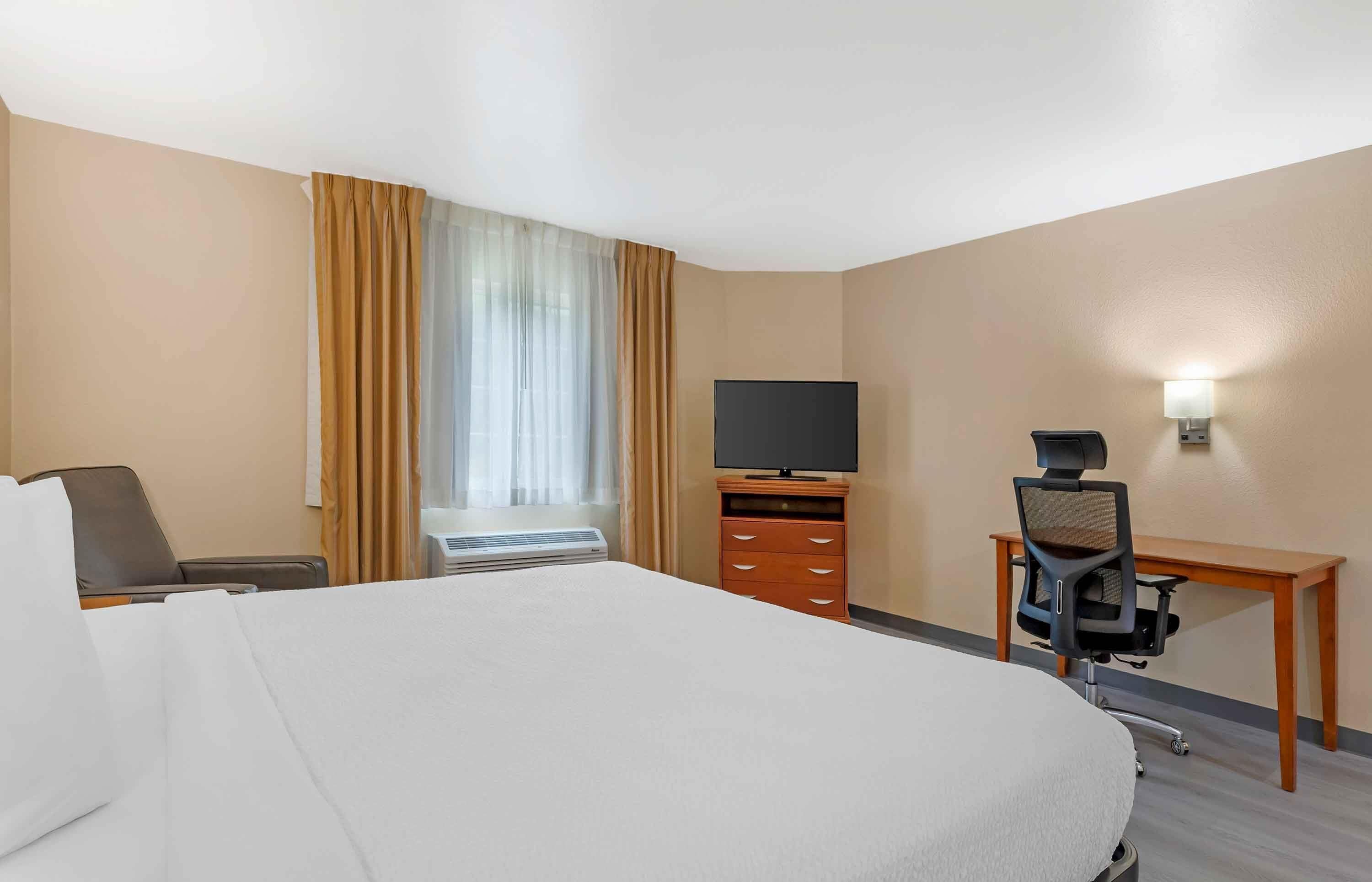 A hotel room with a large bed, a desk with a chair, a television, a dresser, and a window with curtains.
