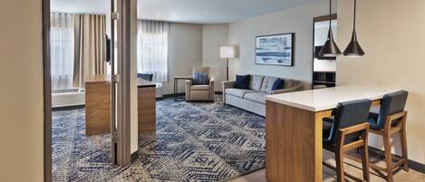 A hotel room with a kitchenette, a sofa, a dining area with bar stools, and a patterned carpet.