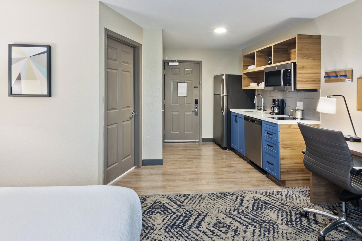 Habitación de hotel compacta con kitchenette, una cama, un escritorio con silla y un tapete estampado.
