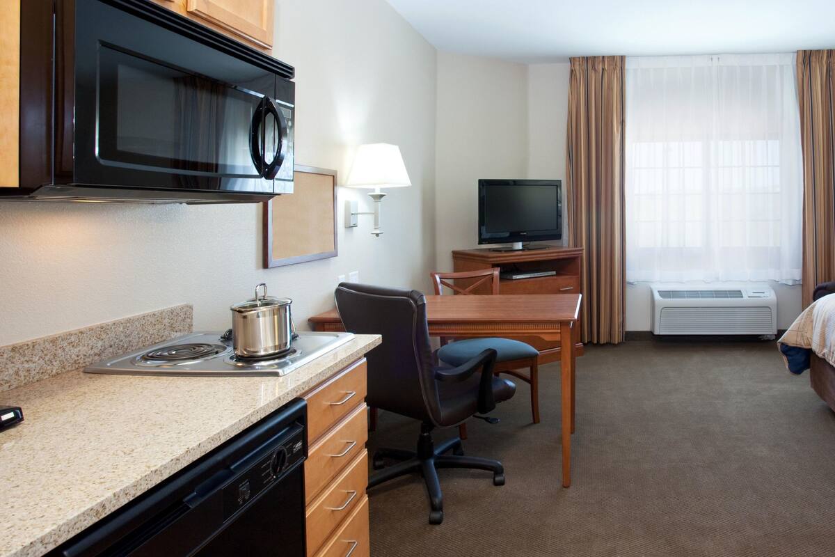 Ein Hotelzimmer mit Küchenzeile, einem Bett, einem Schreibtisch mit Stuhl, einem Fernseher und einem Fenster mit Vorhängen.