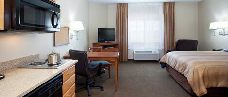A hotel room with a kitchenette, a bed, a desk with a chair, a television, and a window with curtains.