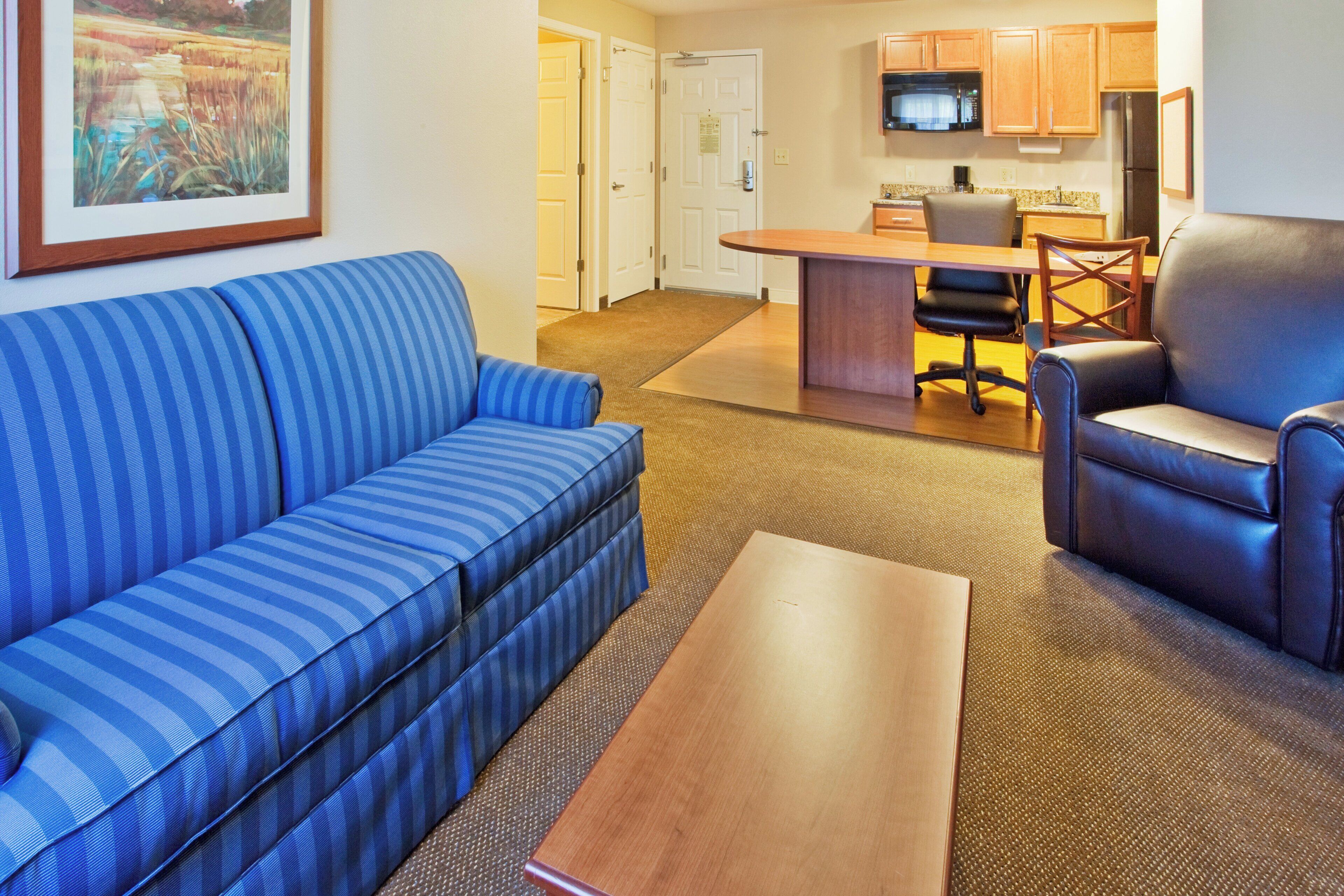 A hotel room with a blue-striped sofa, a wooden coffee table, a black leather chair, and a kitchen area with a microwave.