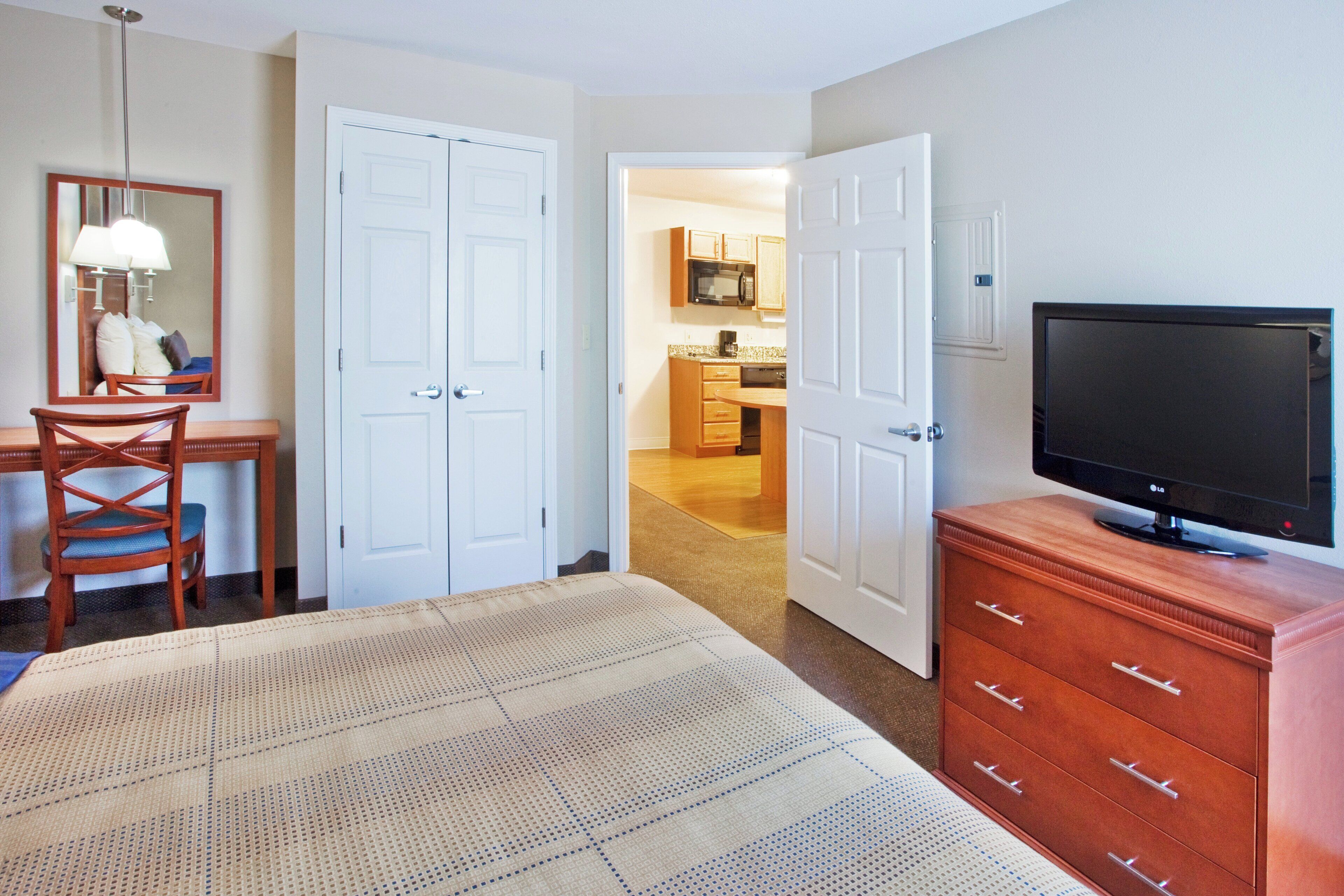 A hotel room with a bed, a television, a dresser, a chair, a desk, and a mirror. There is a hallway leading to a kitchen area.